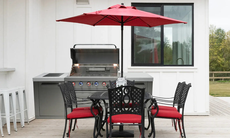 Outdoor patio with a stainless steel grill, red-cushioned black metal chairs, a round table, and a red umbrella. White barstools are in the background.