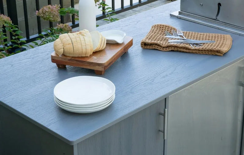 Stack of white plates on a gray outdoor countertop with a wooden cutting board and rolled bread.