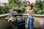 A smiling couple enjoys a barbecue on a sunny terrace, with a grill cooking vegetables and meat. The scene conveys warmth and relaxation.
