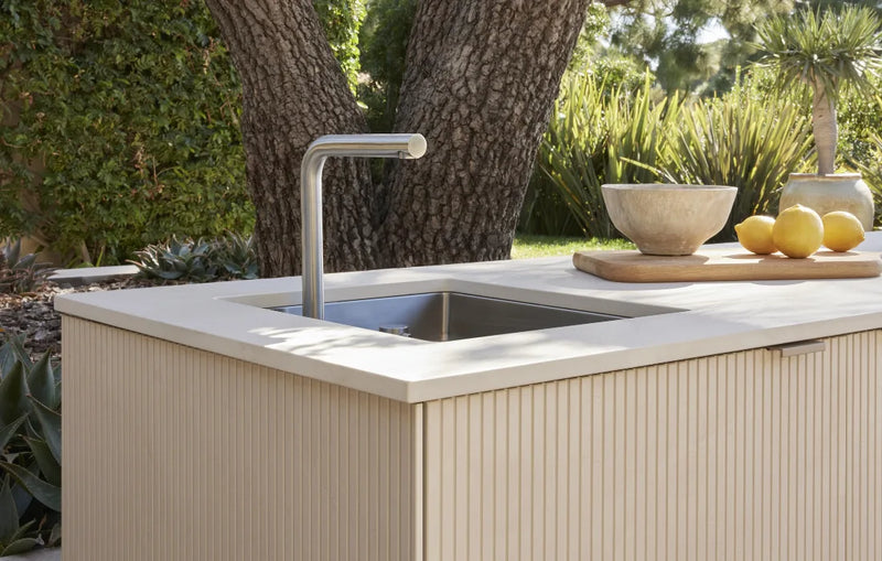 Outdoor kitchen island with a cream ribbed cabinet, a white countertop, a stainless steel sink, and a curved faucet, set in front of a tree with greenery in the background.
