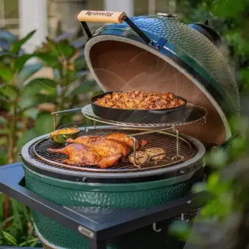 A green ceramic grill with an open lid, showing roasted chicken and a pan of sautéed vegetables. Steam rises, surrounded by green plants, creating a cozy, inviting atmosphere.