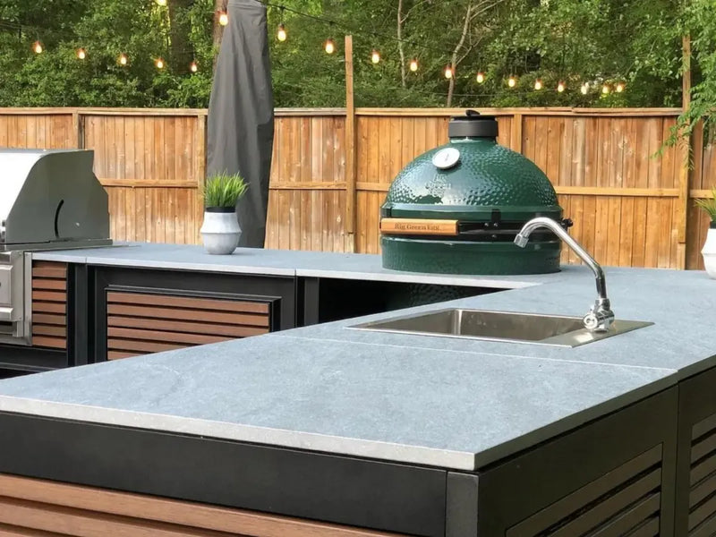 Outdoor kitchen island with a light gray stone countertop, built-in sink, and a green ceramic grill, set against a wooden fence with string lights.