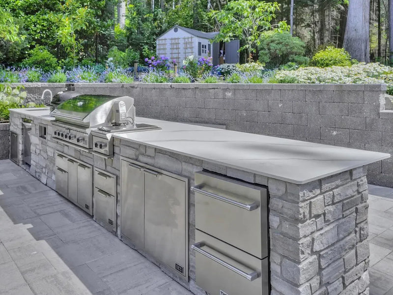 Outdoor kitchen island with a stainless steel gas grill, built-in coolers, and a stone-clad counter on a paved patio with a garden and trees in the background.