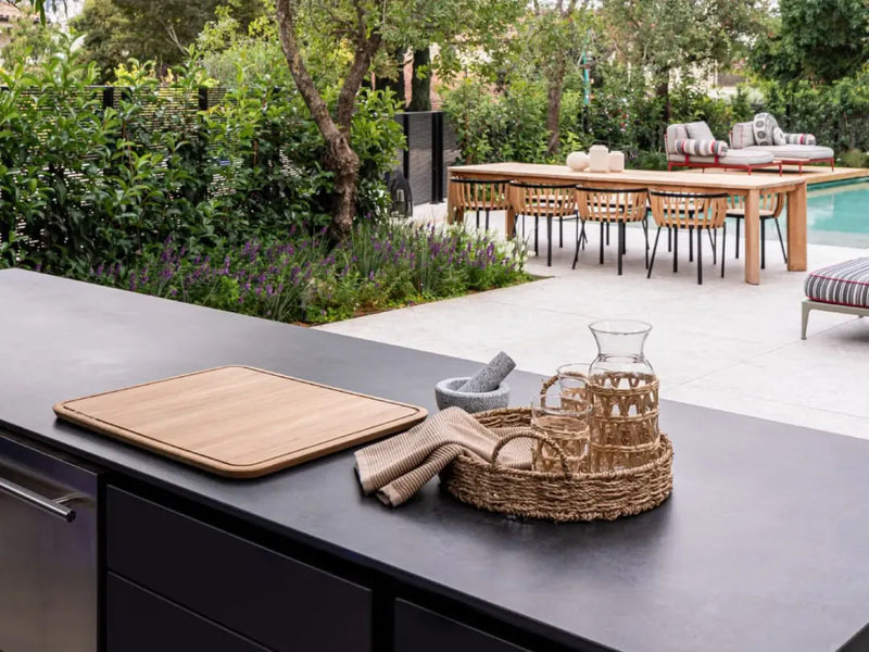 Outdoor kitchen scene with a dark countertop in the foreground holding a wooden cutting board, a folded beige cloth, and a woven tray with a glass carafe and matching glassware. A dining area with a table, chairs, and poolside seating is visible in the background among green plants.