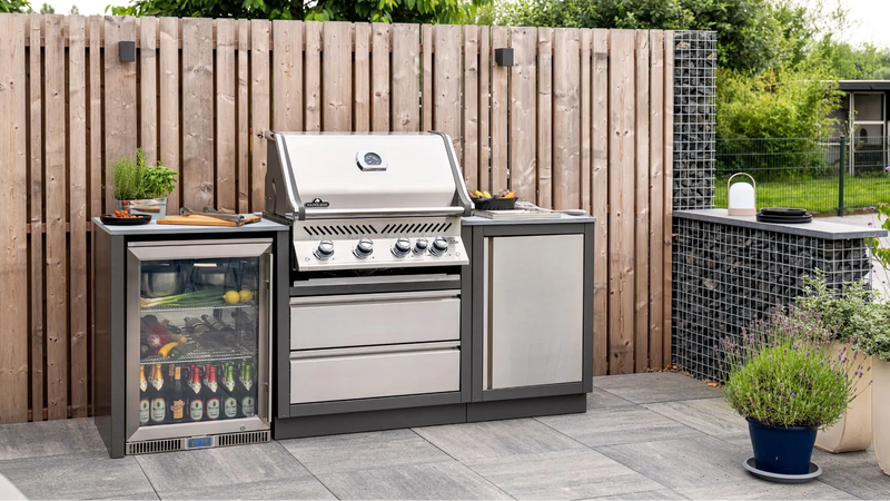 Outdoor kitchen setup with a grill and refrigerator against a wooden fence.