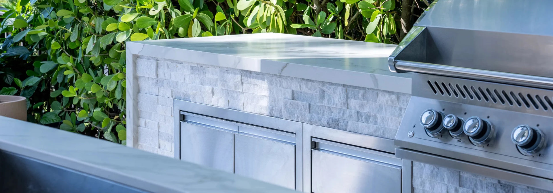 Outdoor stainless steel built-in grill and cooktop unit set into a stone-tile island with green foliage in the background.