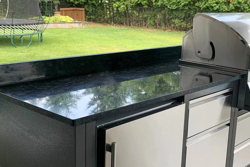 Outdoor kitchen island with a glossy black granite countertop and stainless steel cabinets, shown next to a built-in grill in a backyard setting.