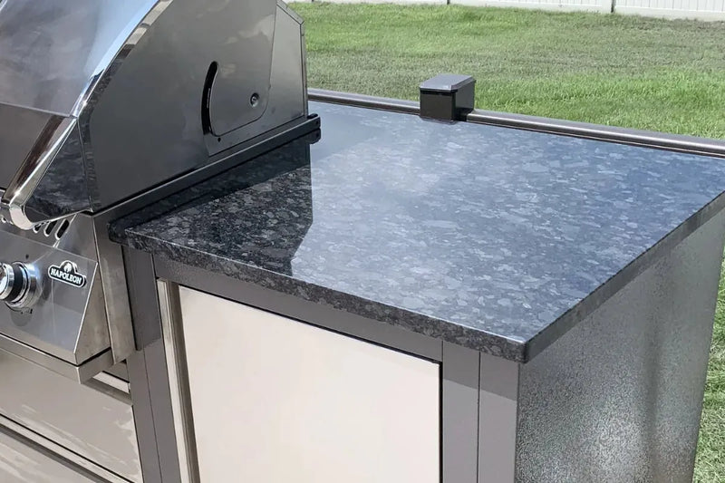 A close-up of an outdoor kitchen setup featuring a dark gray and black speckled granite countertop attached to a stainless steel grill and cabinet base, with a green lawn visible in the background.