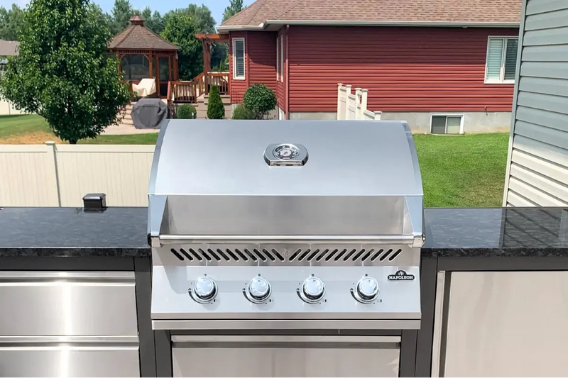 A stainless steel outdoor gas grill with four control knobs and a lid, set into a dark countertop with a backyard view in the background.