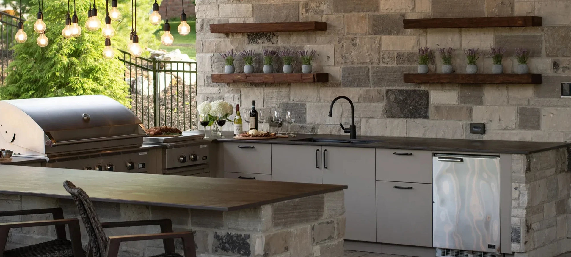 Outdoor kitchen setup with a built-in stainless steel grill, stone tile walls, floating wooden shelves with greenery, a stone-and-wood island, and a stainless steel refrigerator.